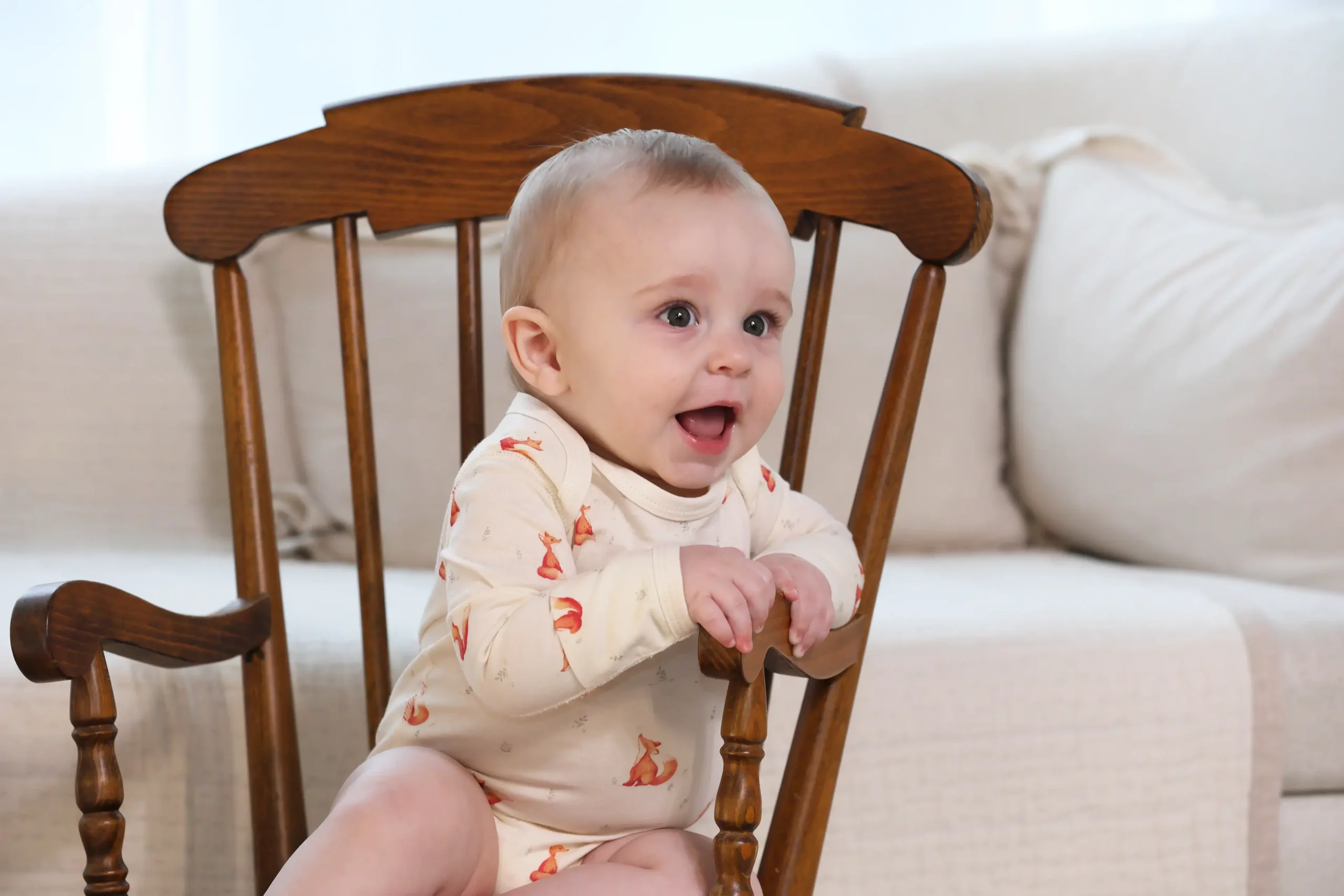 A smiling baby sitting in a wood chair wearing a cotton bodysuit.